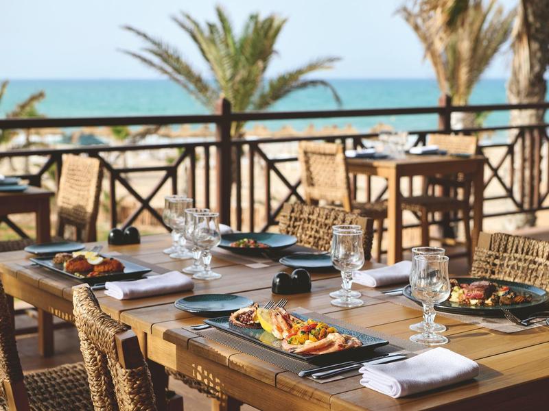 Terraza de un resort junto al mar con mesas puestas, sillas y palmeras al fondo.