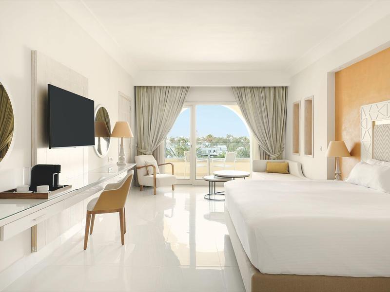 Habitación de hotel luminosa con cama grande, escritorio, TV y vista a terraza y mar.