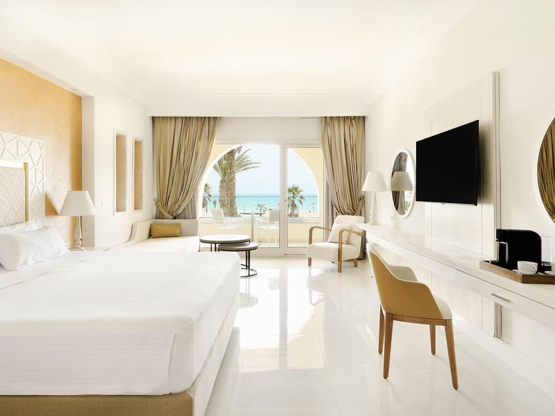 Habitación de hotel luminosa con dos camas, escritorio, silla y vista al mar a través de una gran ventana.