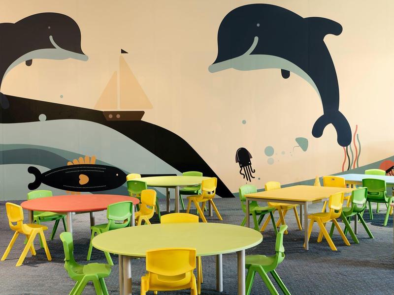 Área de juegos colorida para niños con mesas y sillas pequeñas frente a una pared con decoración de delfines y barcos.