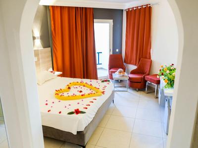 Helles Hotelzimmer mit Bett, herzförmigen Rosenblättern, orangen Vorhängen und Sitzecke.