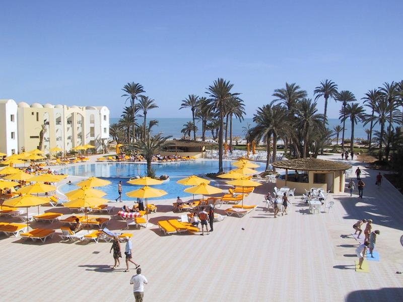 Hotelpoolbereich mit gelben Sonnenschirmen, Palmen und Gästen unter blauem Himmel am Meer.