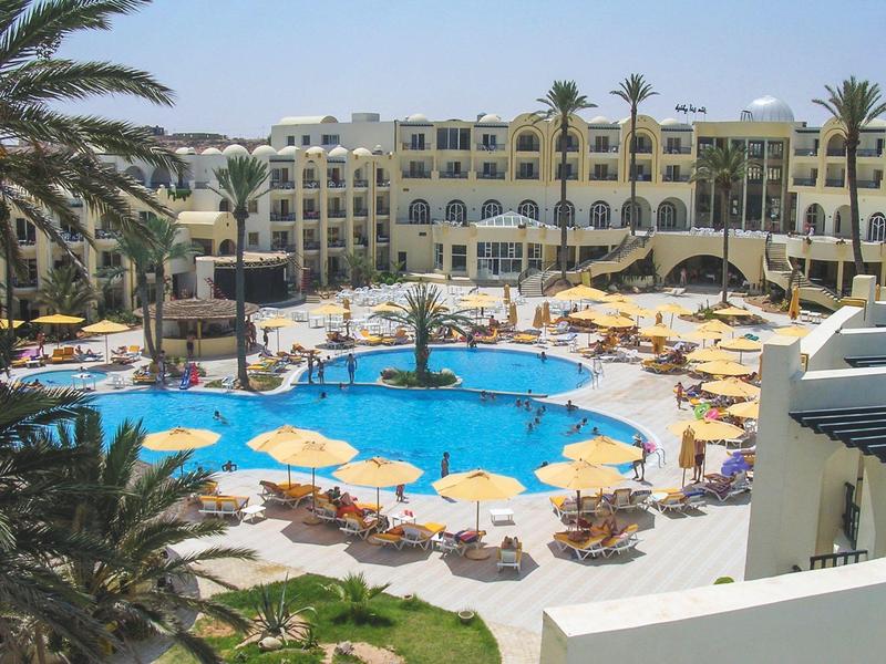 Hotel mit großem Pool, umgeben von Sonnenschirmen und Palmen in sonniger Umgebung.