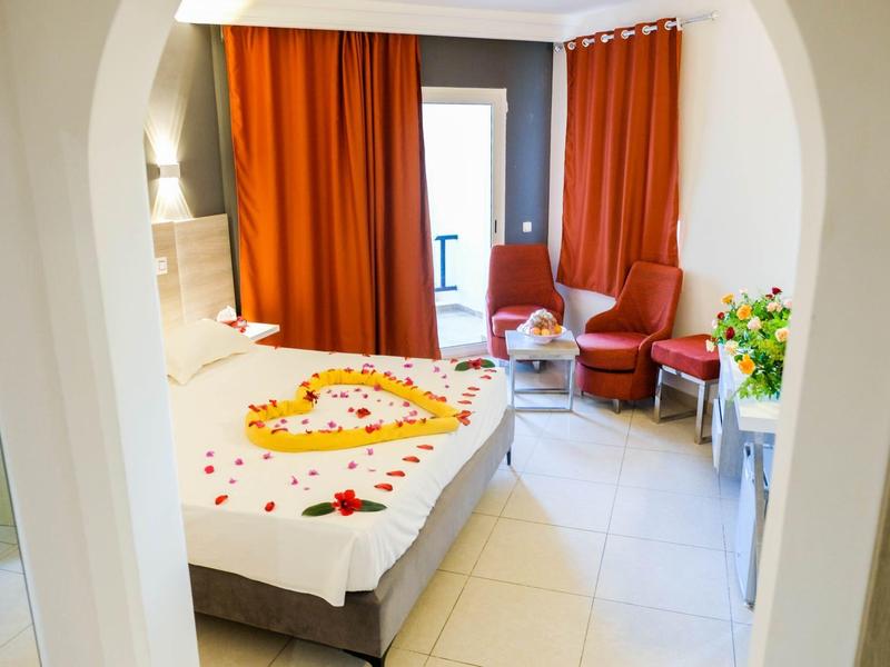 Helles Hotelzimmer mit Bett, herzförmigen Rosenblättern, orangen Vorhängen und Sitzecke.