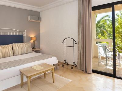 Světelný hotelový pokoj se manželskou postelí, nočním stolkem, stolem, věšákem a balkonem s výhledem na stromy.