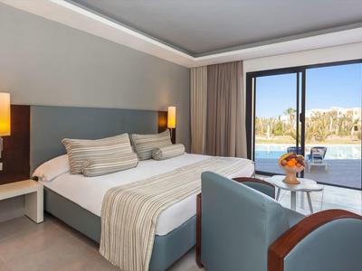 Moderní hotelový pokoj se dvojlůžkem, dvěma nočními stolky, křesly a velkým oknem s výhledem ven.