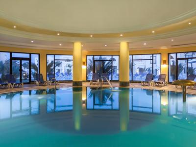 Vnitřní hotelový bazén s velkými okny a žlutými stěnami, obklopený lehátky.