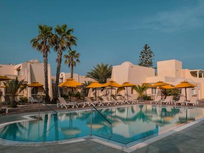 Runder Swimmingpool mit Liegestühlen und gelben Sonnenschirmen, umgeben von Palmen und Gebäuden.