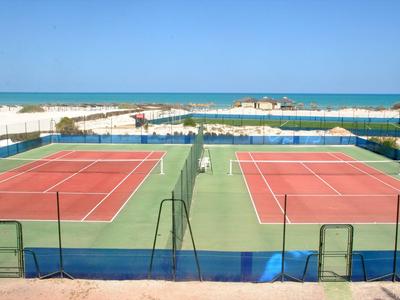 Blick auf zwei rote Tennisplätze am Meer mit blauem Himmel und Sandstrand im Hintergrund.