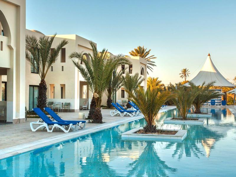 Hotelový bazén s lehátky a palmami při západu slunce.