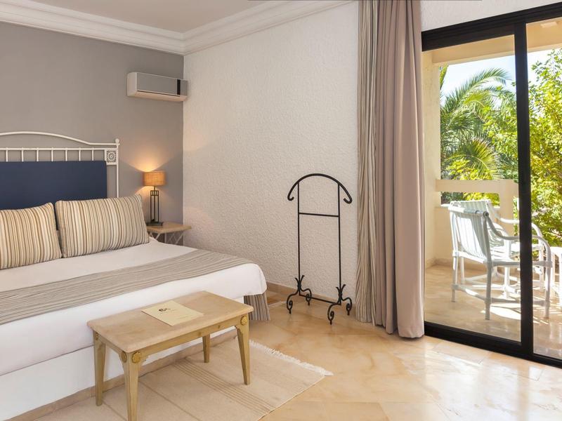 Světelný hotelový pokoj se manželskou postelí, nočním stolkem, stolem, věšákem a balkonem s výhledem na stromy.