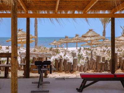 Blick von einer Strandbar mit Fitnessgeräten auf das Meer und Sonnenschirme im Sand.