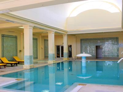 Innenhallen-Schwimmbad mit hellblauen Kacheln, Säulen, Liegestühlen und einem Wasserspilz.