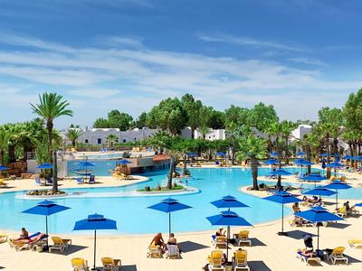 Großer Außenpool mit blauen Sonnenschirmen und Liegen, umgeben von Palmen und weißer Hotelanlage.