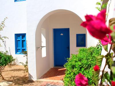 Haus mit weißer Fassade, blauem Fenster und Tür in sonniger Umgebung mit blühenden roten Blumen.