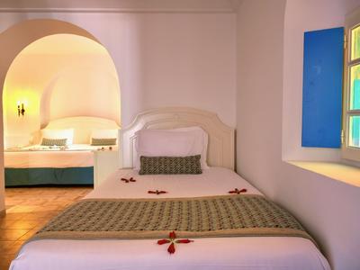 Helles Zimmer mit weißem Bett, grünem Kissen und buntem Fensterlicht, angrenzendem offenen Raum mit Bett.