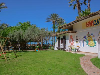 Zielony ogród z huśtawką obok jasnego budynku i palmami w tle.