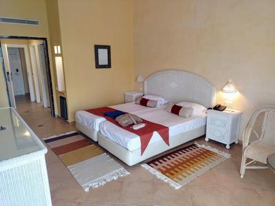 Przytulny pokój hotelowy z podwójnym łóżkiem, stolikami nocnymi i korytarzem z otwartymi drzwiami.