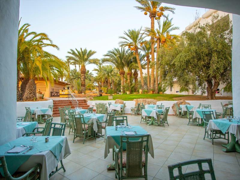 Taras z stołami i krzesłami otoczony palmami i zielenią w ogrodzie hotelowym.