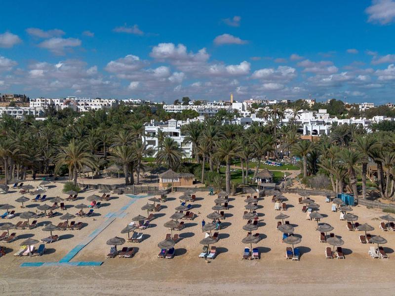 Plaża z leżakami i parasolami w tle miasta z wieloma palmami i białymi budynkami.