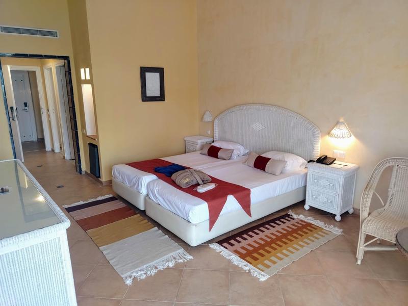 Przytulny pokój hotelowy z podwójnym łóżkiem, stolikami nocnymi i korytarzem z otwartymi drzwiami.