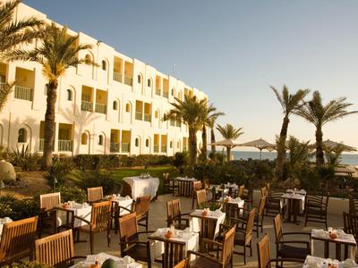 Außenrestaurant mit Tischen und Stühlen vor einem Hotelgebäude mit Palmen und Meerblick bei Sonnenuntergang.