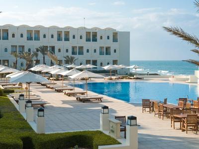 Moderner Hotelpool mit Sonnenliegen und Tischen, Blick auf das Meer bei klarem Himmel.