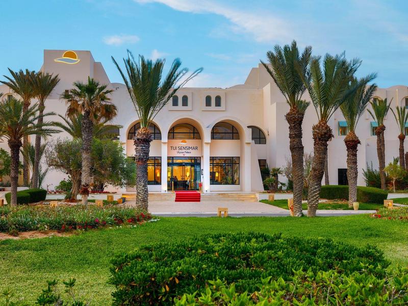 Weißes Gebäude mit Bögen, umgeben von Palmen und grünem Rasen unter blauem Himmel.