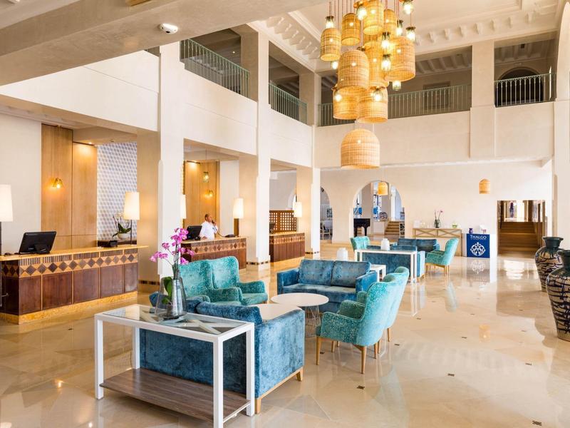 Helle Hotellobby mit modernen Sitzbereichen, hohen Decken und goldenen Pendelleuchten.