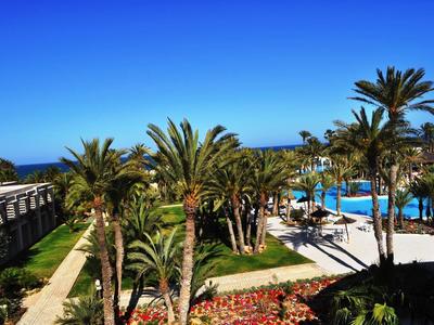 Blick auf einen Poolbereich mit Palmen, blühenden Pflanzen und blauem Himmel am Meer.