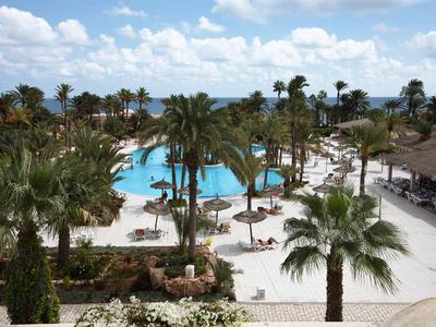 Großer blauer Pool mit Palmen, Liegen und Sonnenschirmen umgeben von weißem Sand und Gebäuden.