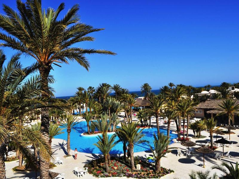 Basen hotelowy otoczony palmami i parasolami pod jasnym, niebieskim niebem.