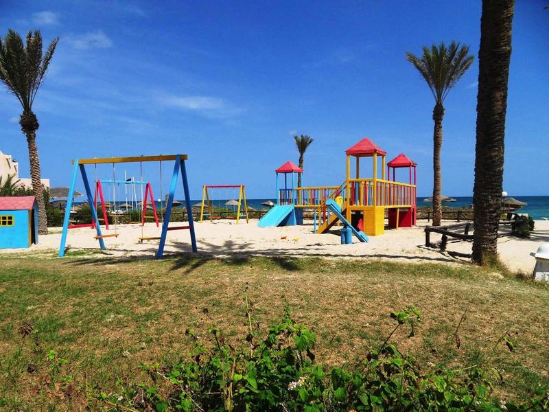 Plac zabaw z huśtawkami i zjeżdżalnią nad morzem, otoczony palmami i trawą.