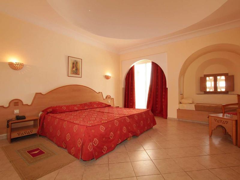 Jasny, bardzo prosto urządzony pokój hotelowy z podwójnym łóżkiem i czerwonawymi zasłonami.