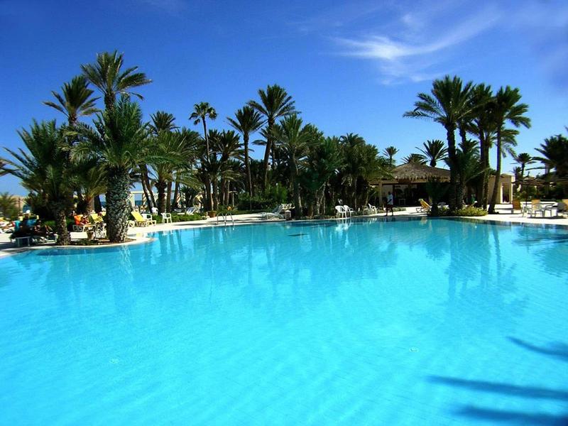 Duży basen z czystą niebieską wodą, otoczony palmami i parasolami pod niebieskim niebem.