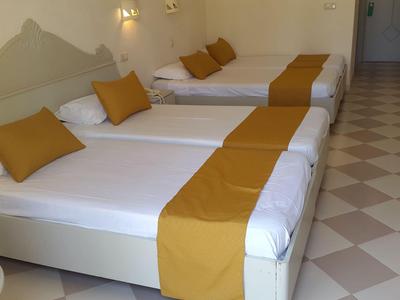 Drei Betten mit weißen Laken und senffarbenen Läufern in einem hellen Hotelzimmer mit Fliesenboden.