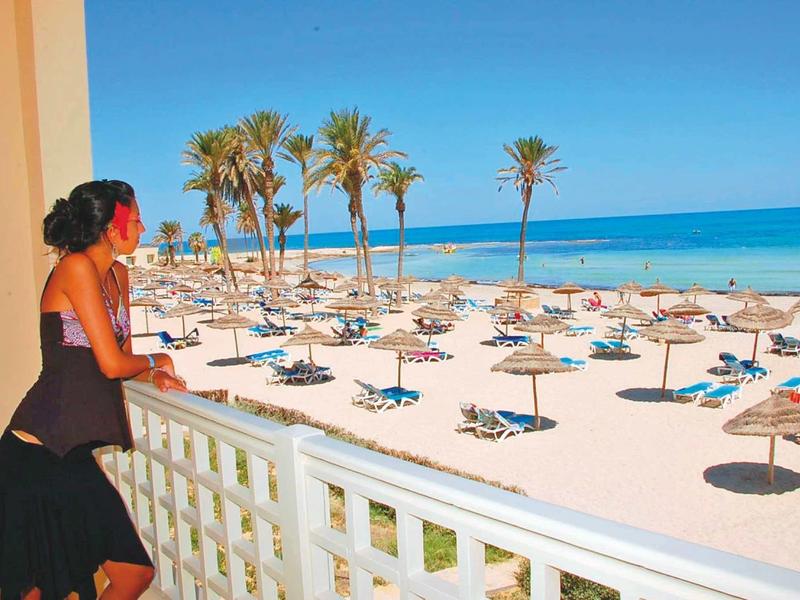 Femme regarde depuis balcon la plage avec parasols et palmiers au bord de la mer.