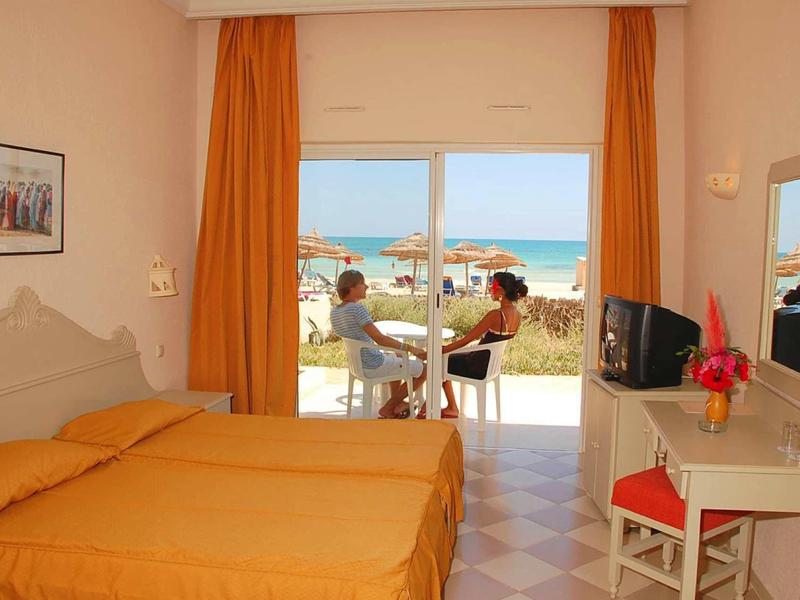 Chambre d'hôtel avec deux lits, coin salon sur balcon avec vue sur la mer, rideaux orange.