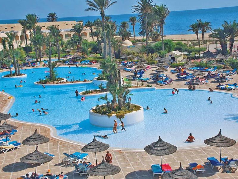 Grand piscine d'hôtel avec palmiers, chaises longues et vue sur la mer par temps ensoleillé.