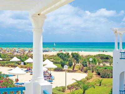 Blick von Terrasse mit weißen Säulen auf grünen Garten, Strand und blaues Meer unter wolkigem Himmel.