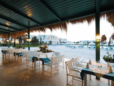 Offenes Strandrestaurant mit gedeckten Tischen, weißen Stühlen und Blick auf Meer und Boote.