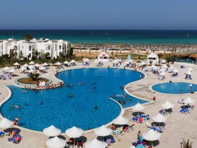 Großer blauer Pool mit weißen Sonnenschirmen und umliegenden weißen Gebäuden am Meer.