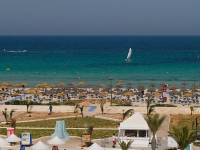 Strand mit vielen Sonnenschirmen, Menschen, Palmen und blauem Meer mit Segelboot und klarem Himmel.