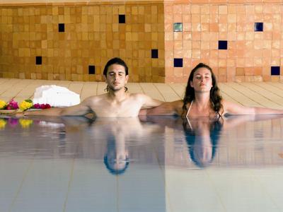 Zwei entspannte Personen in einem Schwimmbecken vor einer orangefarbenen Wand mit dunklen Fliesen.