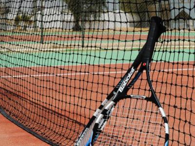 Tennisschläger lehnt am Netz, zwei gelbe Tennisbälle liegen auf rotem Spielfeld.