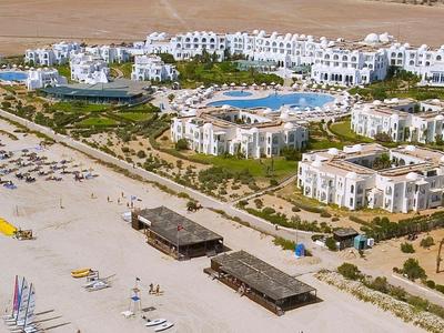 Luftaufnahme eines Resorts mit Pool, weißen Gebäuden und einem Sandstrand mit Sonnenliegen.