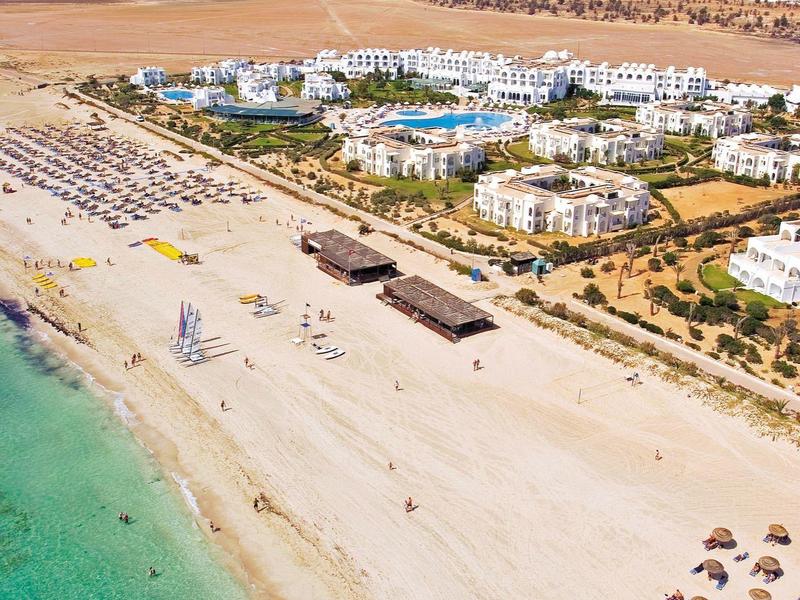 Písečná pláž s tyrkysovým mořem, hotely a slunečníky pod modrou oblohou.