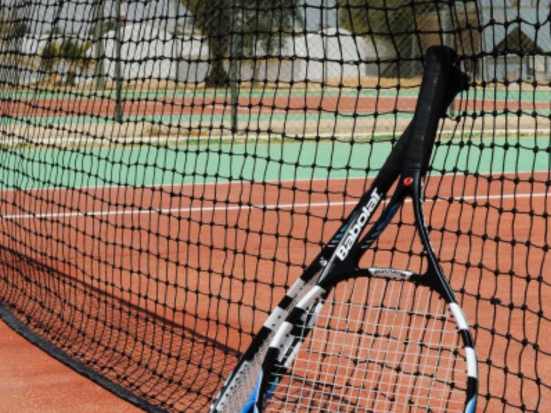 Tenisová raketa opřená o síť, dvě žluté míčky na červeném venkovním kurtu.