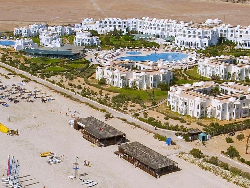 Luftaufnahme eines Resorts mit Pool, weißen Gebäuden und einem Sandstrand mit Sonnenliegen.