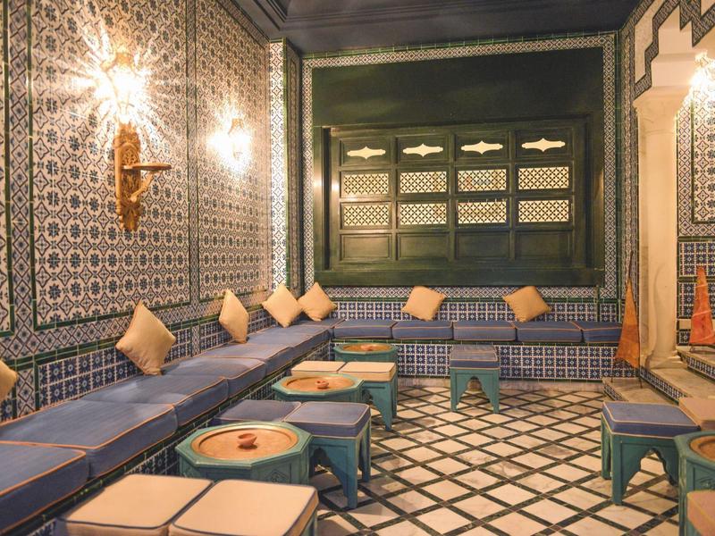 Marocký salon s modrými lavicemi, béžovými polštáři a zdobenými dlaždicovými stěnami.